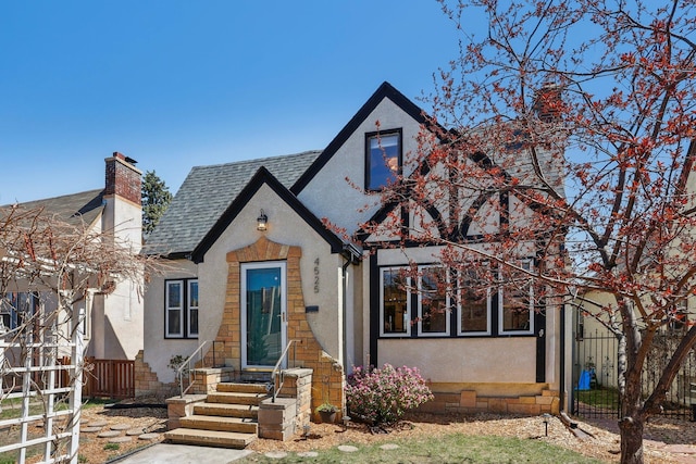 tudor-style house featuring fence, stucco siding, a chimney, and roof with shingles