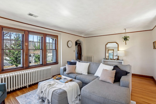 living room featuring visible vents, arched walkways, light wood-style floors, radiator, and ornamental molding