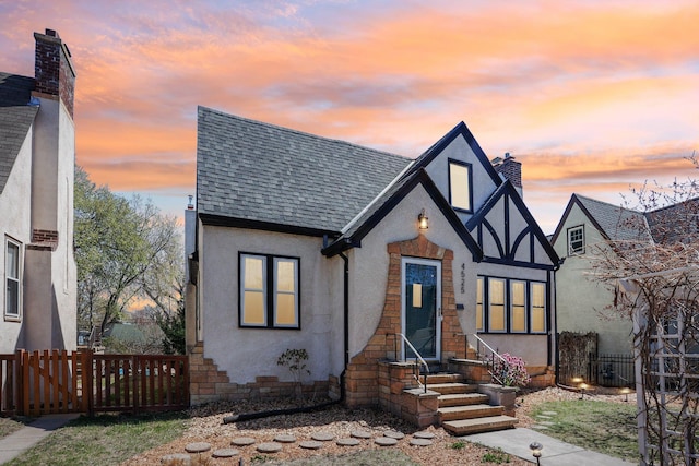 tudor home with fence, roof with shingles, and stucco siding