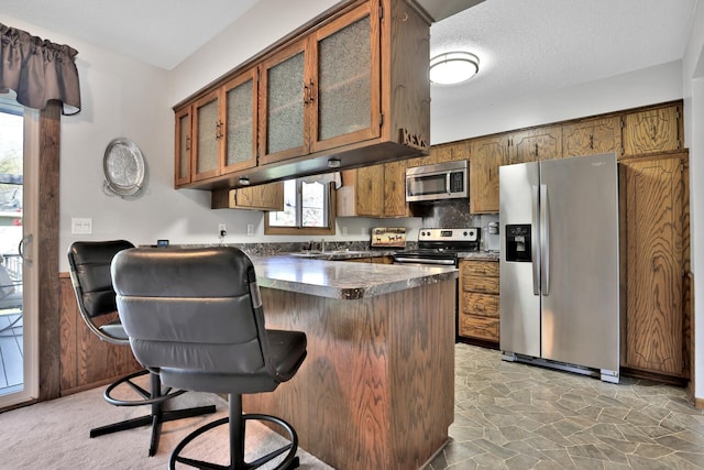 kitchen featuring stone finish floor, a peninsula, brown cabinets, dark countertops, and appliances with stainless steel finishes