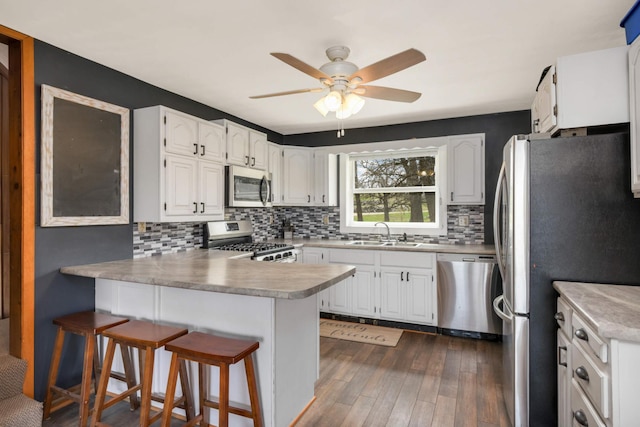 kitchen featuring stainless steel appliances, a peninsula, a sink, backsplash, and dark wood-type flooring