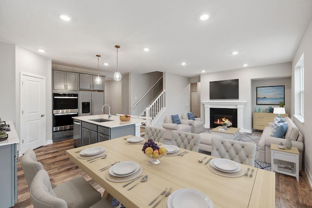dining area with a warm lit fireplace, dark wood-style flooring, stairs, and recessed lighting