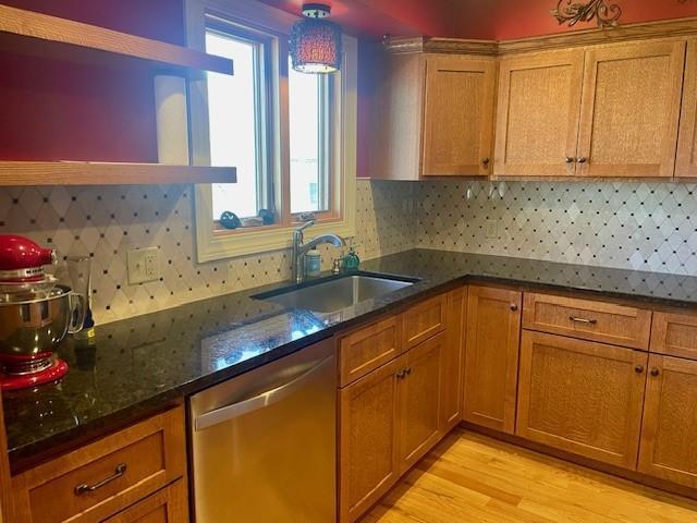 kitchen featuring stainless steel dishwasher, a sink, brown cabinets, backsplash, and dark stone countertops