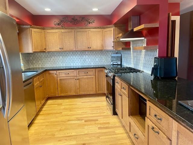 kitchen with stainless steel appliances, open shelves, light wood finished floors, wall chimney exhaust hood, and dark stone countertops