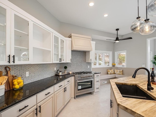 kitchen featuring custom exhaust hood, decorative backsplash, stainless steel stove, wood counters, and a sink