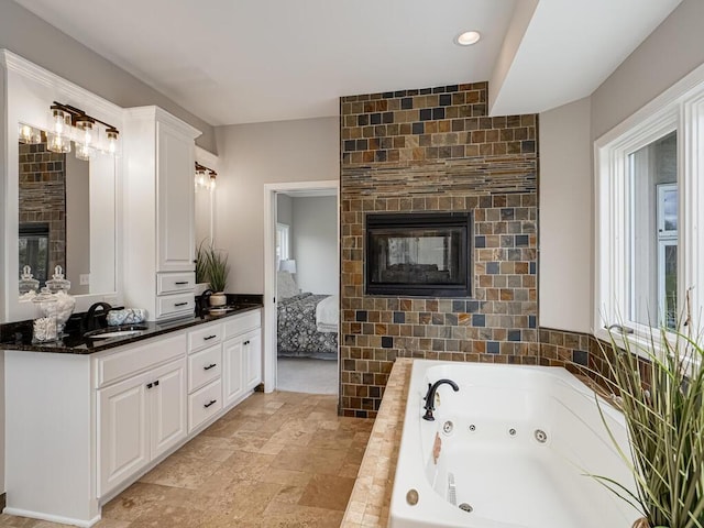 full bathroom with a jetted tub, a multi sided fireplace, stone finish floor, and vanity