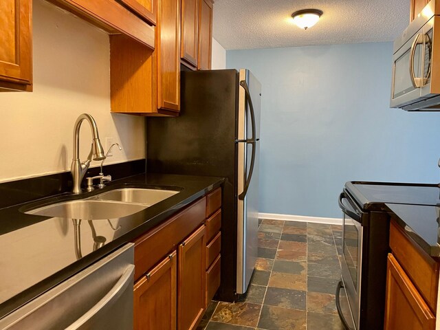 kitchen with stainless steel appliances, a sink, brown cabinetry, a textured ceiling, and dark countertops
