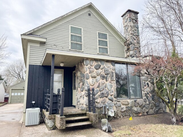 view of front facade featuring stone siding, central AC unit, a chimney, and an outbuilding