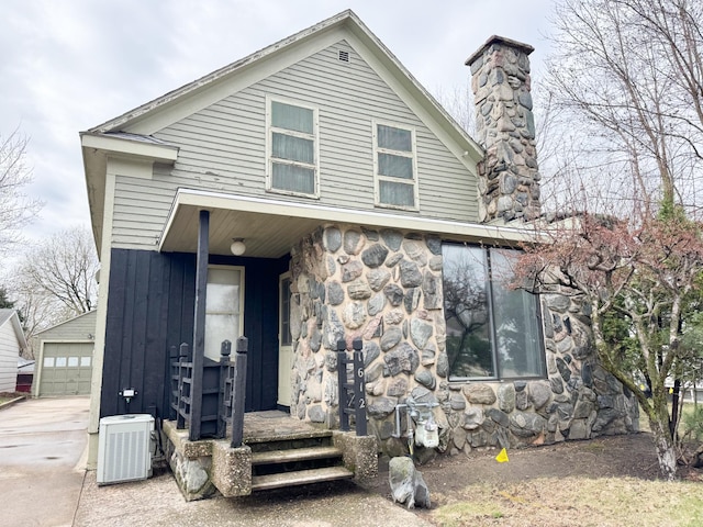 view of front of property featuring stone siding, central air condition unit, a chimney, and an outbuilding