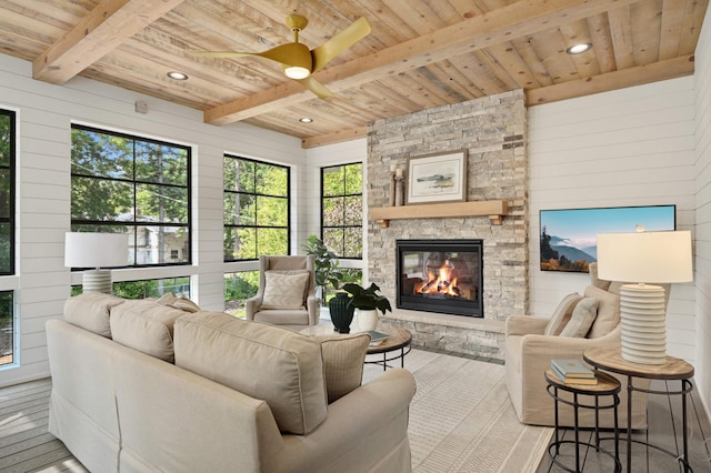 living area with beamed ceiling, ceiling fan, wooden ceiling, wooden walls, and a stone fireplace
