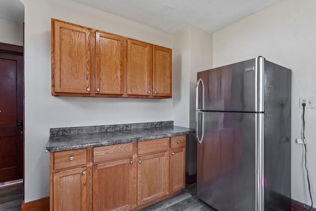 kitchen featuring brown cabinetry, dark countertops, dark wood-type flooring, and freestanding refrigerator