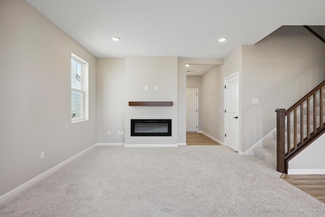 unfurnished living room featuring a glass covered fireplace, stairway, carpet floors, and baseboards