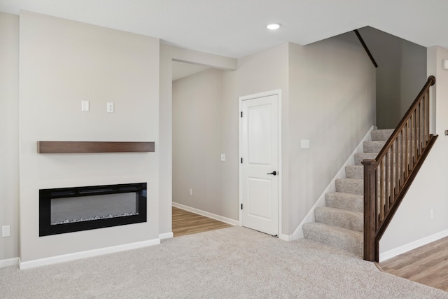 unfurnished living room featuring a glass covered fireplace, stairway, carpet, and baseboards