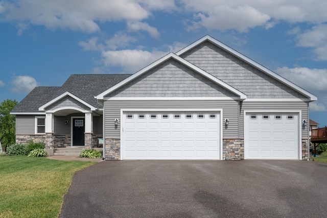 craftsman-style house with driveway, stone siding, roof with shingles, a front yard, and an attached garage