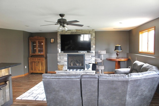 living area featuring baseboards, ceiling fan, dark wood-style flooring, and a stone fireplace