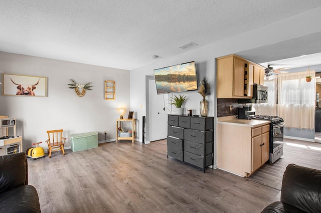 kitchen featuring a textured ceiling, stainless steel microwave, light brown cabinetry, gas stove, and wood finished floors