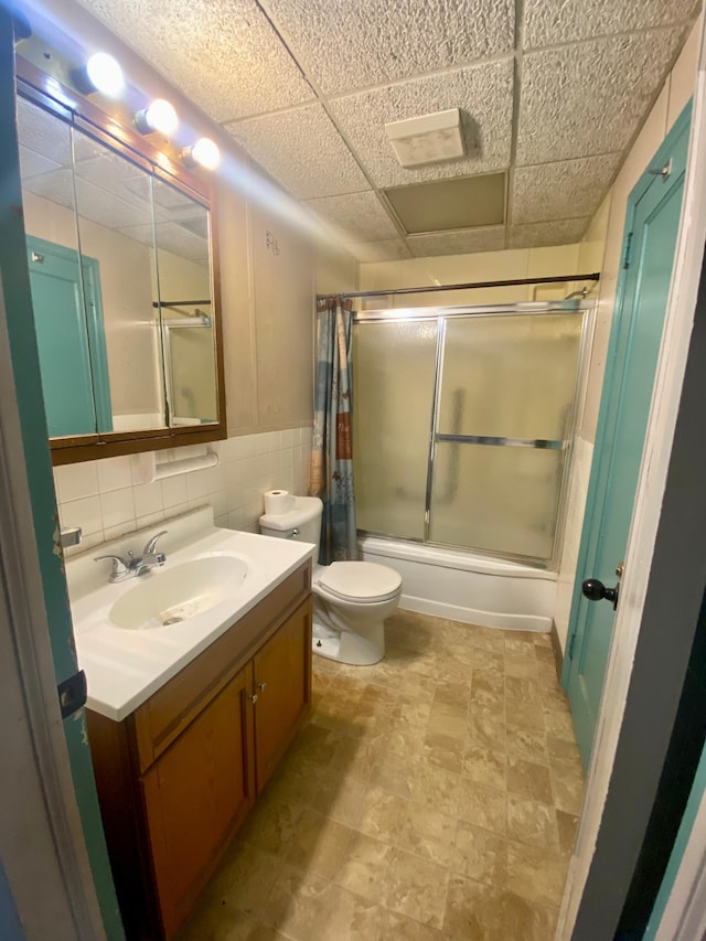 full bath with shower / bath combination with glass door, a drop ceiling, toilet, tile walls, and vanity