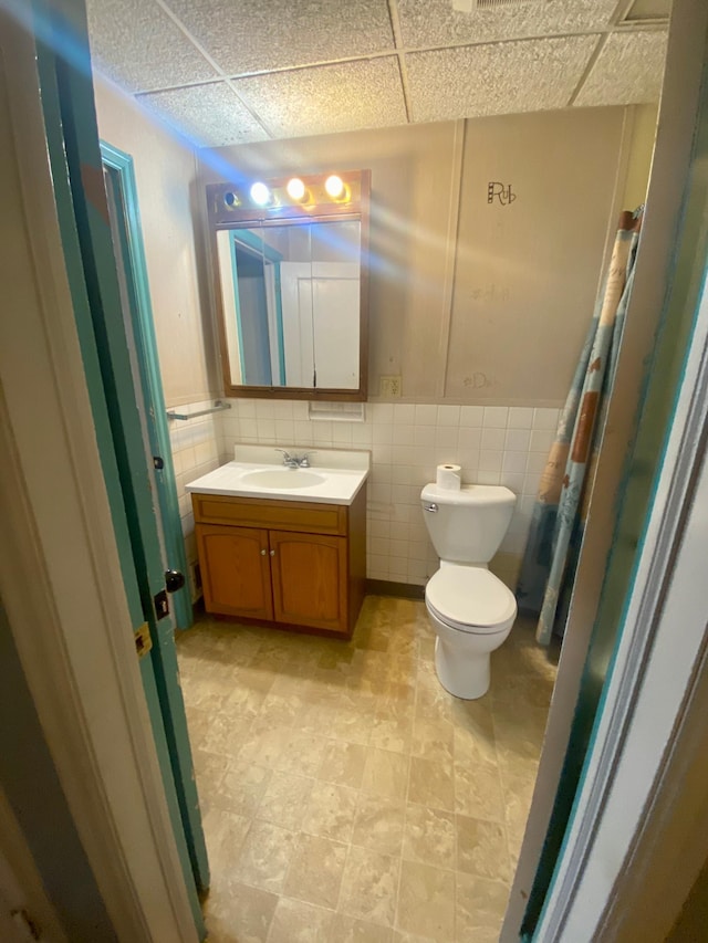 bathroom with a drop ceiling, a shower with curtain, tile walls, toilet, and vanity