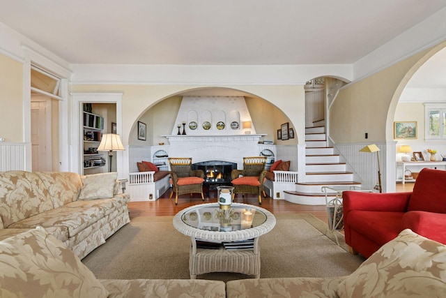 living room with arched walkways, a fireplace, wood finished floors, stairway, and a wainscoted wall