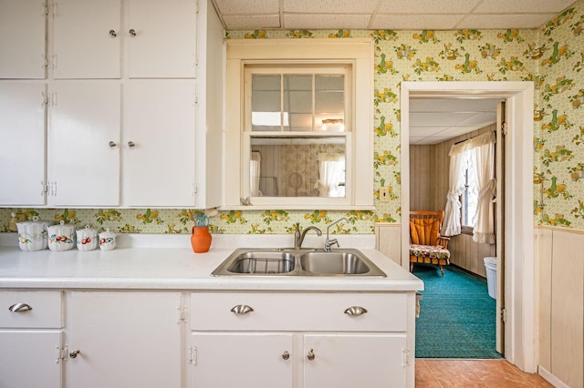 kitchen with a sink, light countertops, white cabinetry, wallpapered walls, and a drop ceiling