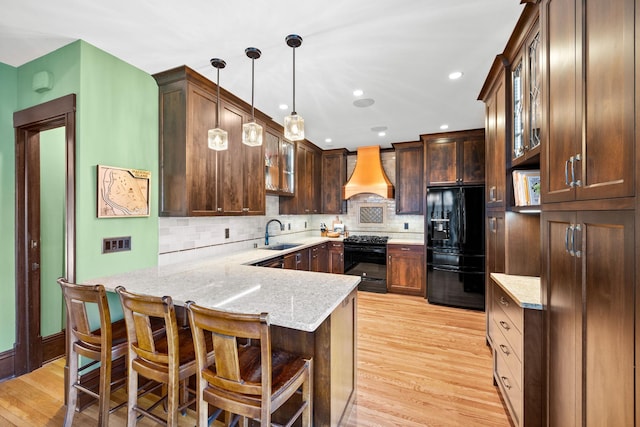 kitchen with a sink, black appliances, a peninsula, custom range hood, and light wood-style flooring