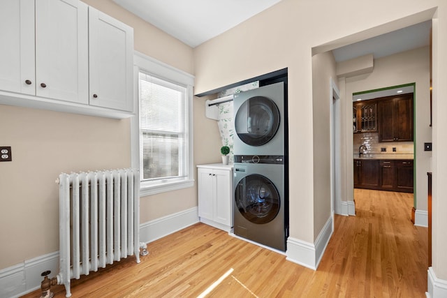 laundry area with radiator heating unit, baseboards, stacked washer and clothes dryer, and light wood-type flooring