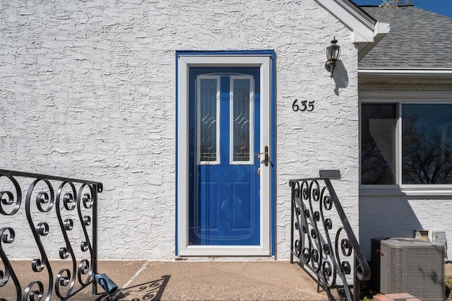 property entrance with stucco siding, a shingled roof, and central air condition unit