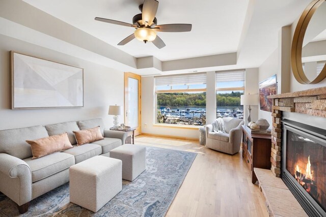 living room featuring ceiling fan, a raised ceiling, a brick fireplace, and light wood-style floors