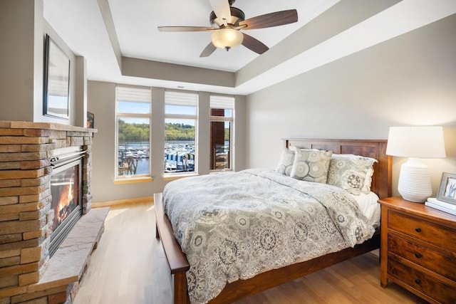 bedroom featuring light wood-style flooring, a raised ceiling, ceiling fan, and a glass covered fireplace