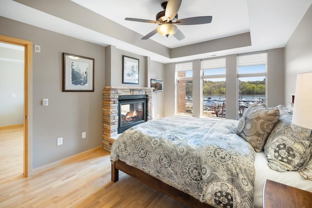 bedroom with a ceiling fan, a tray ceiling, light wood-style flooring, baseboards, and a fireplace