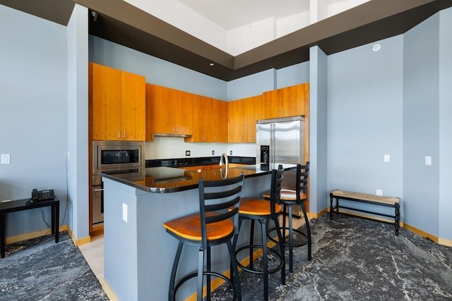 kitchen with dark countertops, stainless steel microwave, brown cabinetry, and an island with sink