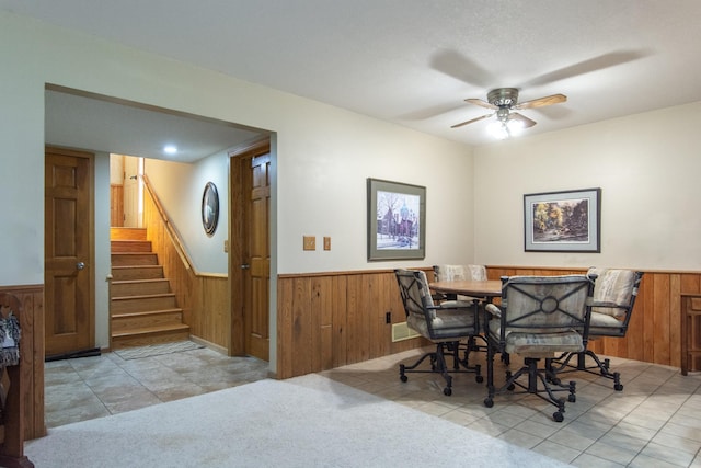 tiled dining space featuring stairs, a ceiling fan, wood walls, and wainscoting