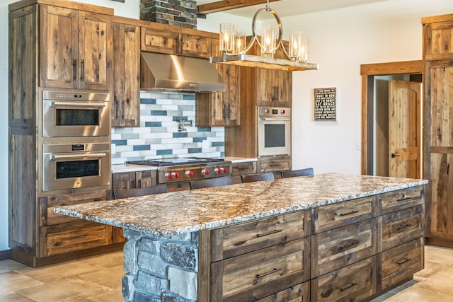 kitchen with under cabinet range hood, light stone counters, hanging light fixtures, backsplash, and stainless steel appliances