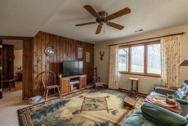living room with carpet floors, a textured ceiling, visible vents, a ceiling fan, and wooden walls