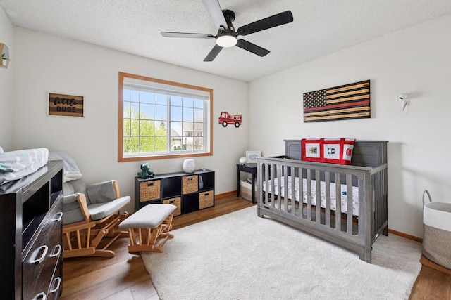 bedroom with a ceiling fan, a textured ceiling, a nursery area, wood finished floors, and baseboards