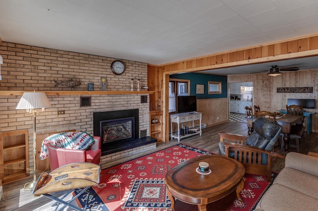 living room featuring wooden walls, wood finished floors, a brick fireplace, and a ceiling fan