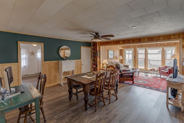 dining space featuring wood walls, ceiling fan, a baseboard heating unit, wainscoting, and wood finished floors