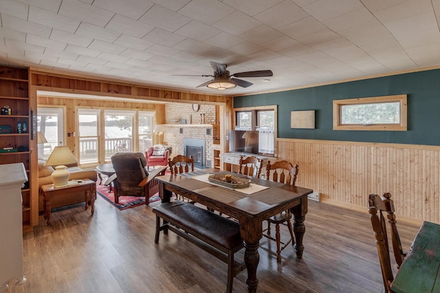 dining room featuring wood walls, a brick fireplace, ceiling fan, a wainscoted wall, and wood finished floors