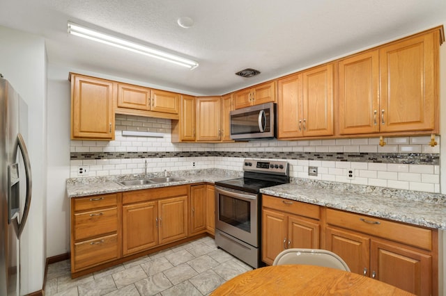 kitchen with appliances with stainless steel finishes, a sink, backsplash, light stone counters, and a textured ceiling