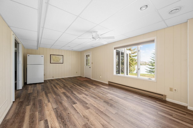 unfurnished living room with a baseboard heating unit, dark wood-type flooring, a ceiling fan, a paneled ceiling, and baseboards