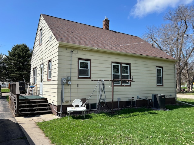 rear view of property featuring a chimney, cooling unit, a yard, and roof with shingles