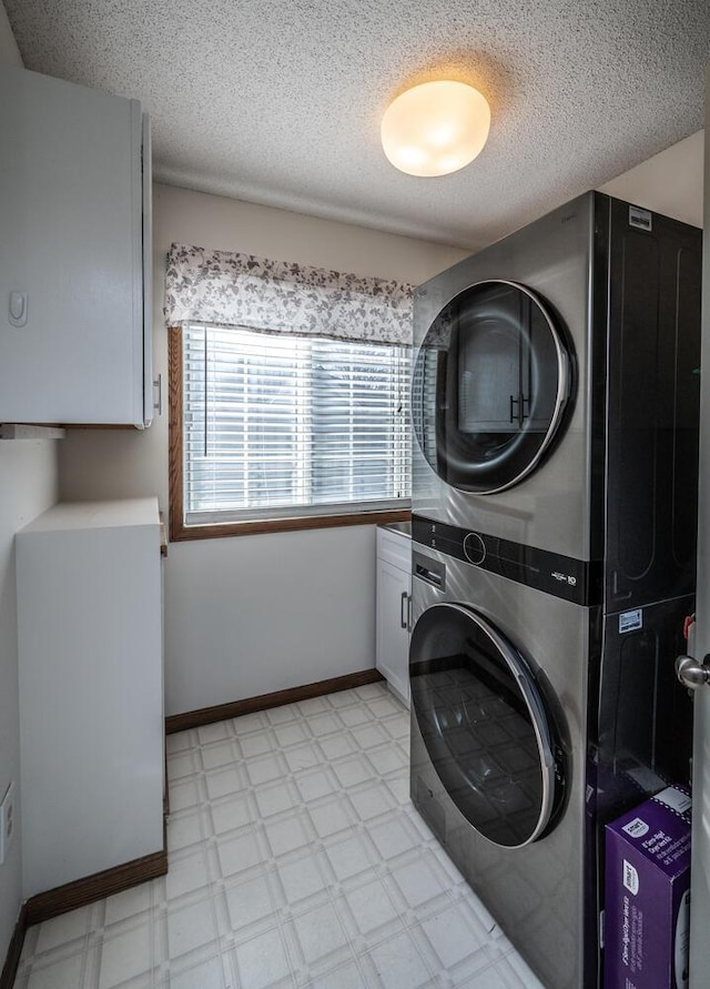 laundry room featuring stacked washer / drying machine, cabinet space, light floors, a textured ceiling, and baseboards