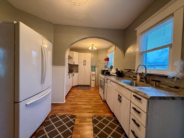 kitchen with dark countertops, a sink, white appliances, arched walkways, and wood finished floors