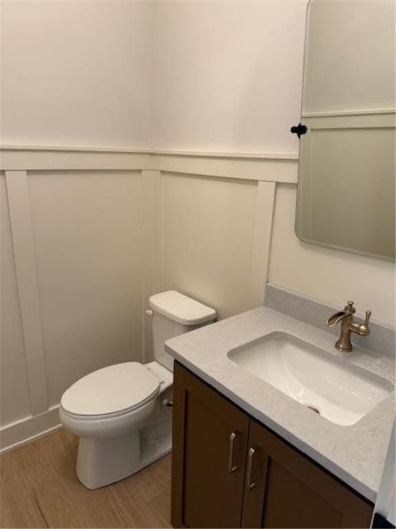 half bathroom featuring a decorative wall, toilet, wood finished floors, vanity, and wainscoting