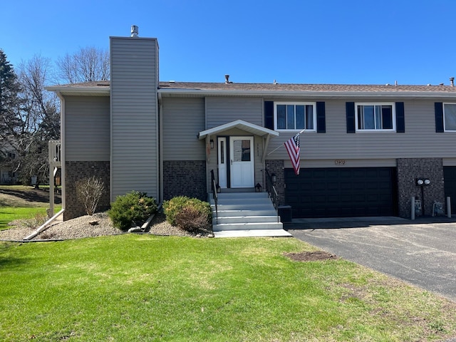 bi-level home with aphalt driveway, a front yard, a chimney, brick siding, and an attached garage