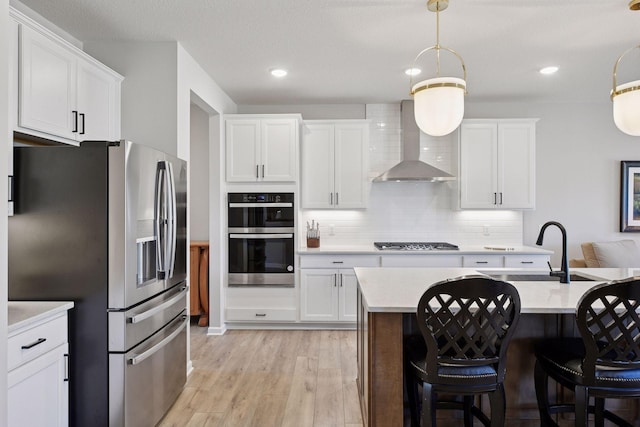 kitchen featuring decorative backsplash, appliances with stainless steel finishes, wall chimney exhaust hood, light countertops, and light wood-style flooring