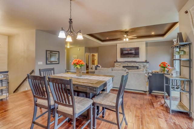 dining area with a stone fireplace, a raised ceiling, light wood finished floors, and a ceiling fan