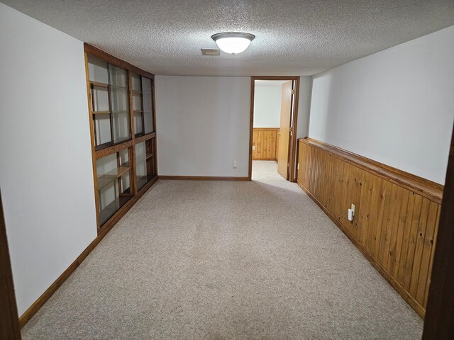 empty room featuring wooden walls, light colored carpet, a wainscoted wall, a textured ceiling, and baseboards
