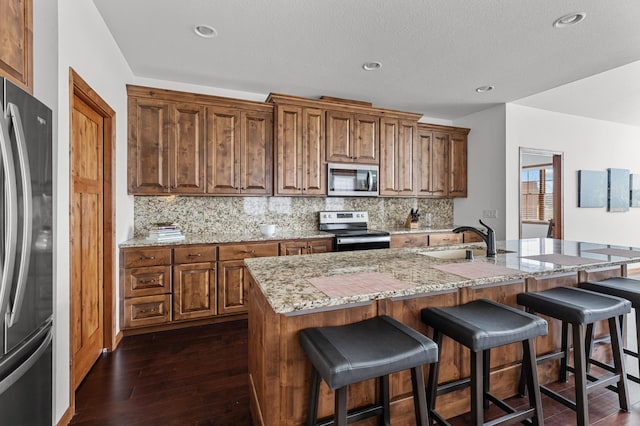 kitchen with brown cabinetry, backsplash, appliances with stainless steel finishes, light stone countertops, and a sink