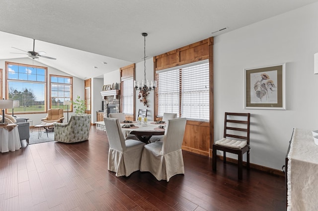 dining area featuring dark wood-style flooring, a stone fireplace, baseboards, ceiling fan with notable chandelier, and vaulted ceiling